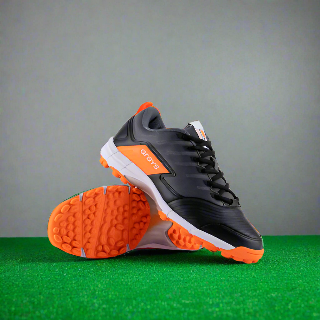 A pair of Grays Flash Junior Astro hockey shoes with a black upper, orange accents, and a rubber outsole visible in an orange hue.