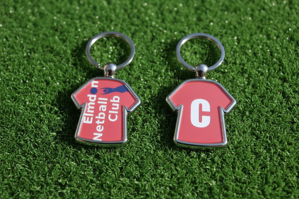 Two small metal keychains shaped like red sports jerseys are shown side by side on a light wood surface. The left keychain has a ring and a jersey with the text 'Elmdon Netball Club' in white and blue.