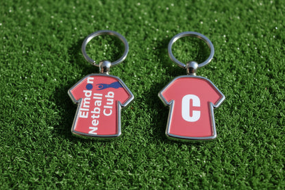 Two small metal keychains shaped like red sports jerseys are shown side by side on a light wood surface. The left keychain has a ring and a jersey with the text 'Elmdon Netball Club' in white and blue.