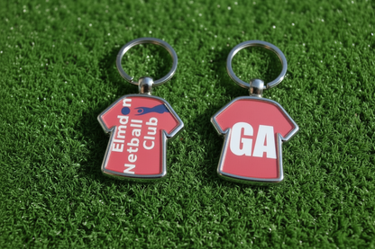 Two red football shirt keychains with silver metal rings, displayed on a green artificial turf background. One keychain features the text 'Elmden Netball Club' in white with a blue diagonal stripe, and the other features the initials 'GK' in white.