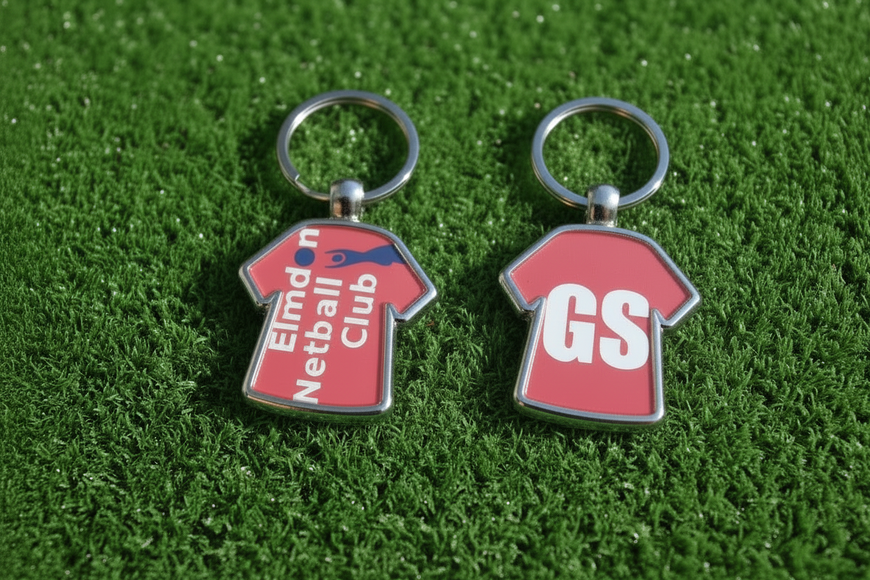 Two custom metal keychains shaped like red sports jerseys on a green grass background. The left keychain features a red shirt with 'Elmdon Netball Club' in white text and a blue diagonal stripe.