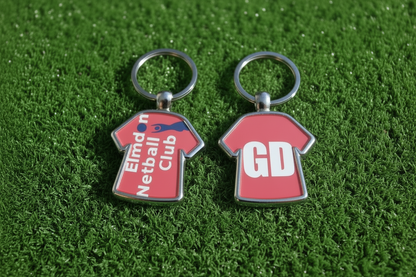 Two red football shirt keychains with silver metal rings, displayed on a green artificial turf background. One keychain features the text 'Elmdon Netball Club' in white with a blue netball graphic, and the other features the initials 'GD' in white.