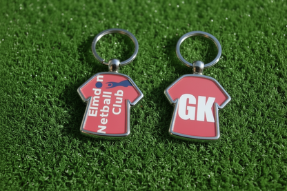 Two red football shirt keychains with silver metal rings, displayed on a green artificial turf background. One keychain features the text 'Elmdon Netball Club' in white with a blue diagonal stripe, and the other features the initials 'GK' in white.